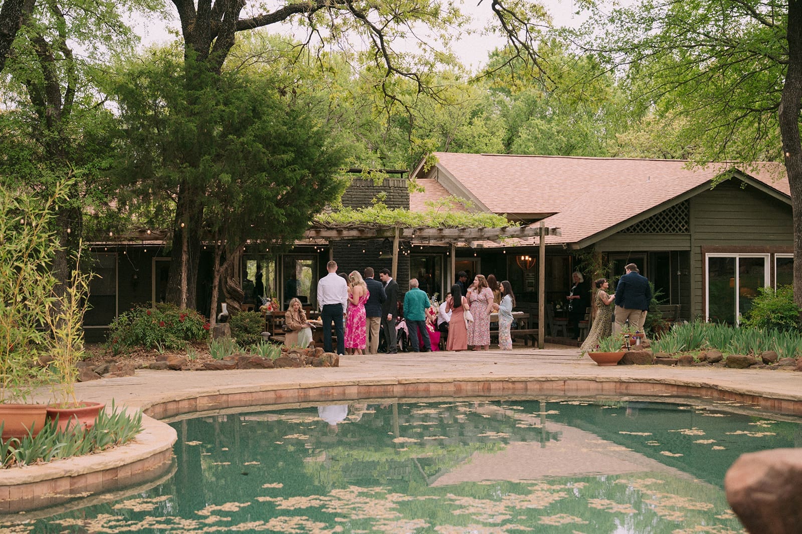 A group of people stands and talks near a house with a covered porch, surrounded by trees and a pool with leaves floating on the water.