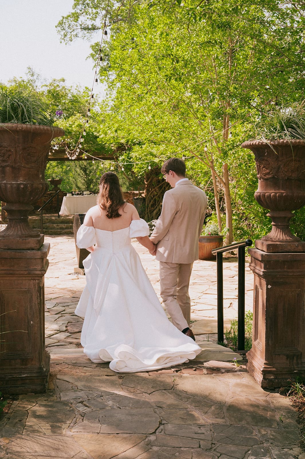 Bride and groom walk along a brick path surrounded by greenery, with the bride wearing a long white veil and holding a bouquet for a Weston Gardens Wedding