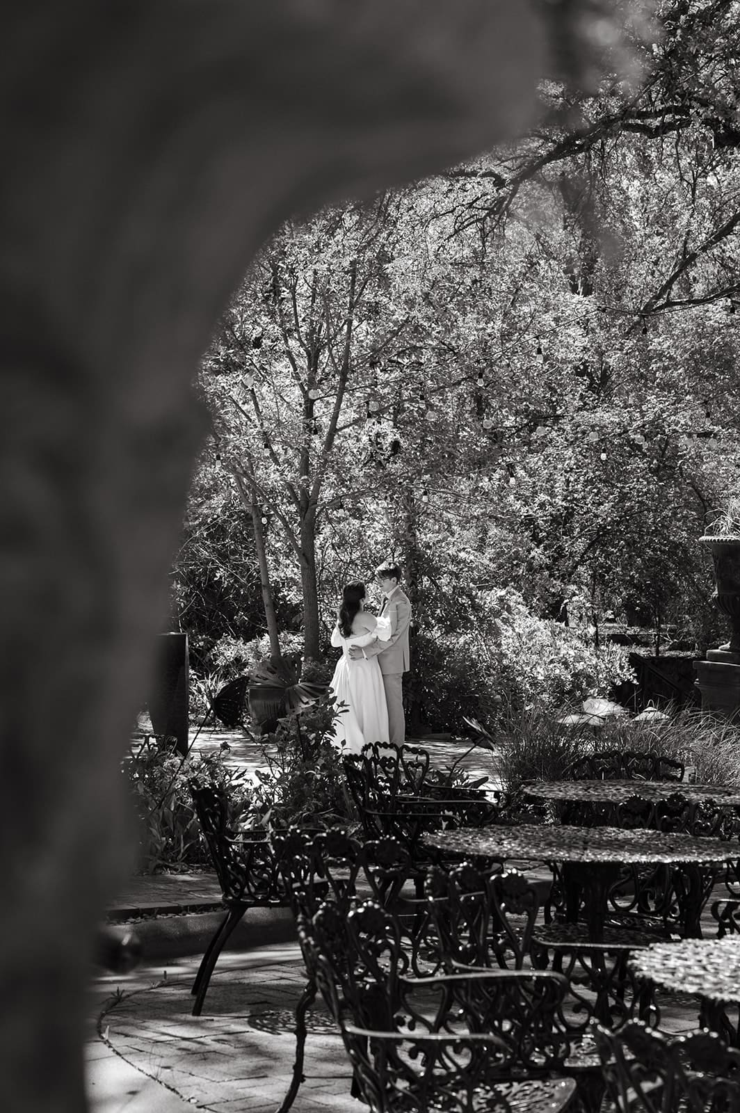 A black and white photo shows a person in white standing among trees in a garden, with empty metal tables and chairs in the foreground.