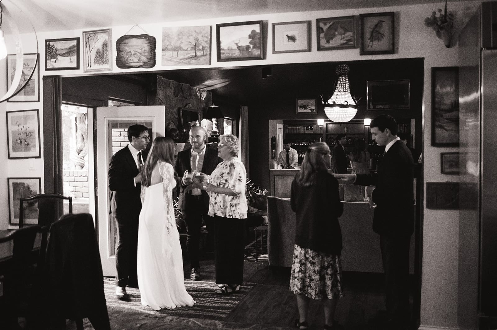 A small group of people, including a bride and groom, gather and converse in a room decorated with framed pictures and art on the walls.