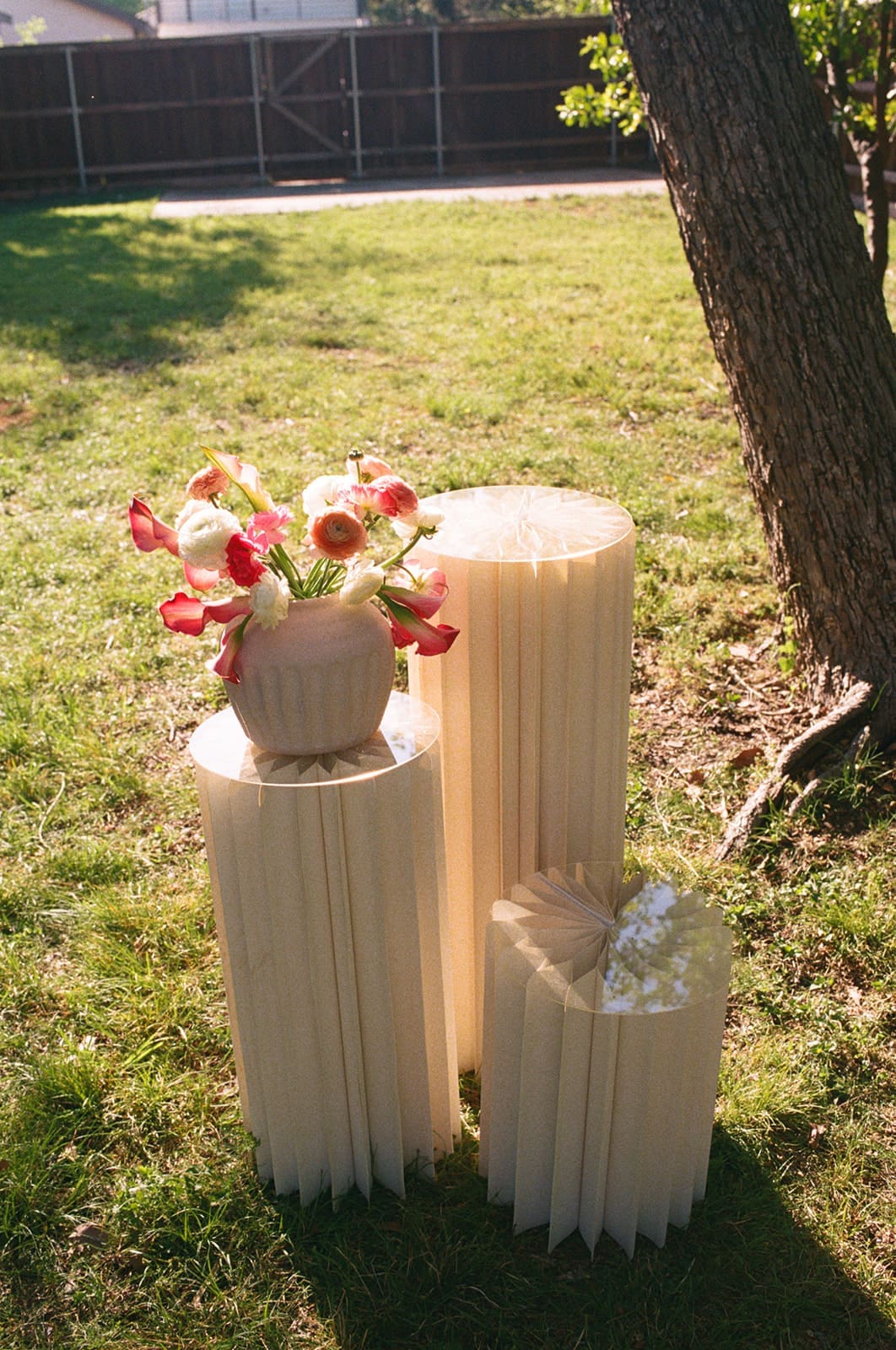 Three cream-colored, pleated pedestal stands of varying heights stand on grass, with a vase of red and white flowers placed on the tallest stand.