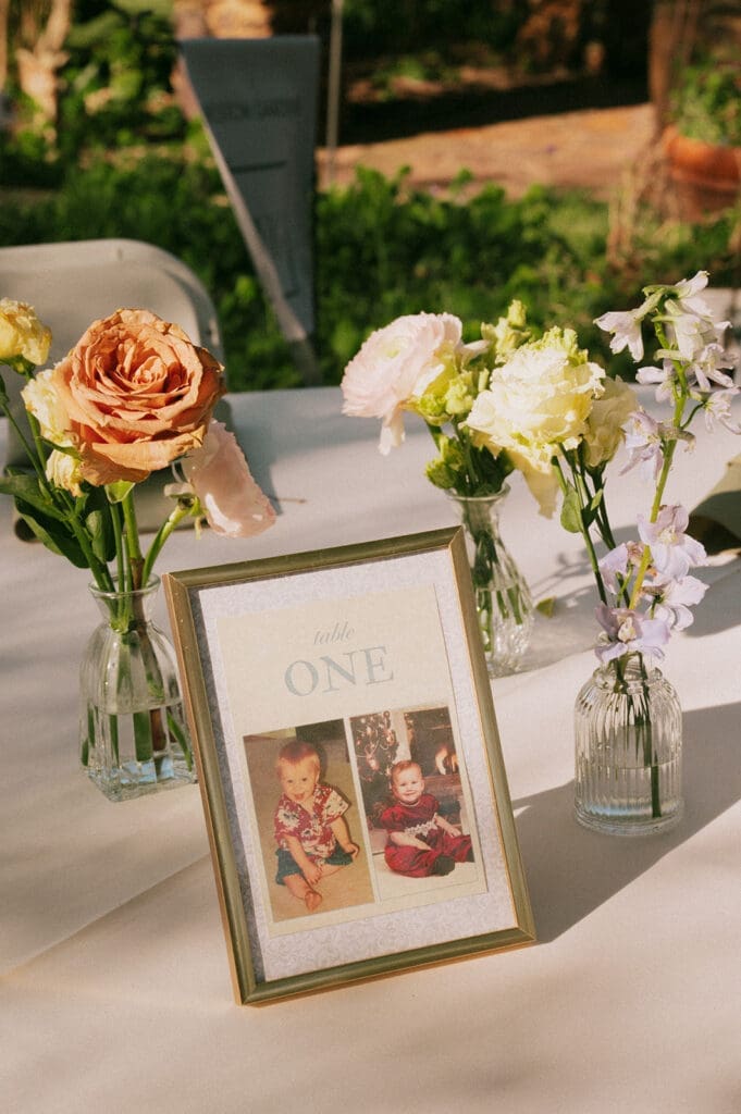 A framed photo display labeled "ONE" sits on a table with small vases holding assorted roses and flowers, outdoors in natural light for a Weston Gardens Wedding
