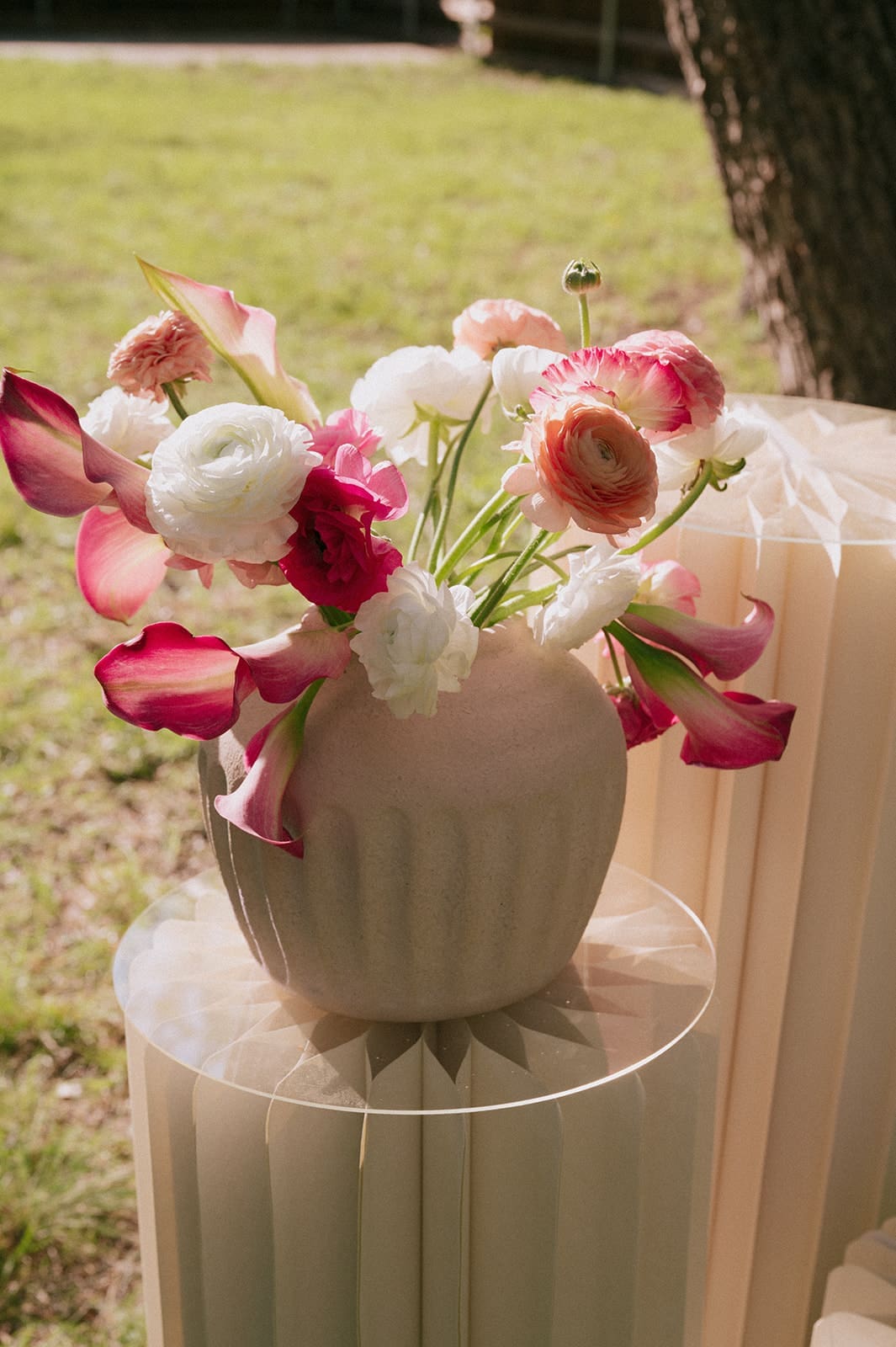 Three cream-colored, pleated pedestal stands of varying heights stand on grass, with a vase of red and white flowers placed on the tallest stand.