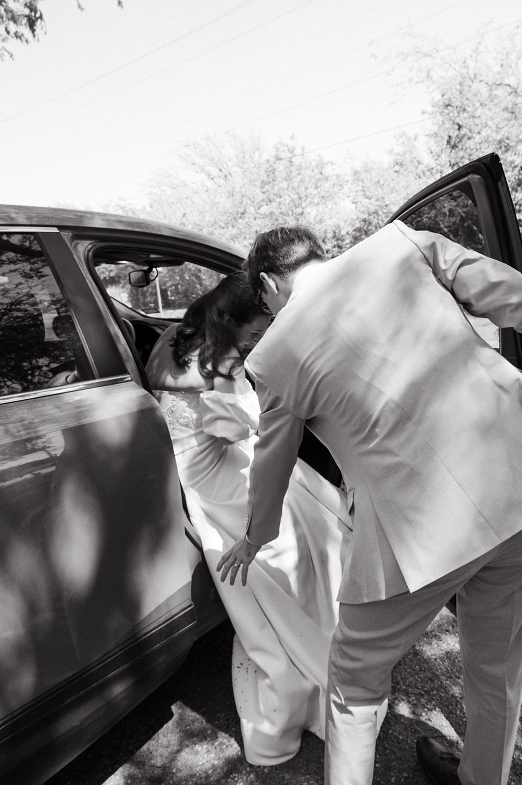 A bride in a wedding dress is helped out of a car by a man in a suit, photographed in black and white.
