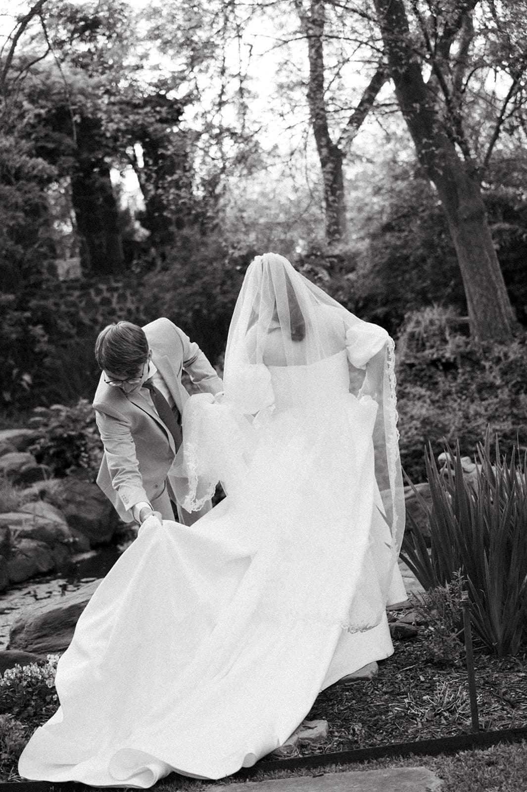 A groom assists a bride with her long wedding dress as they stand outdoors in a garden setting for a Weston Gardens Wedding 