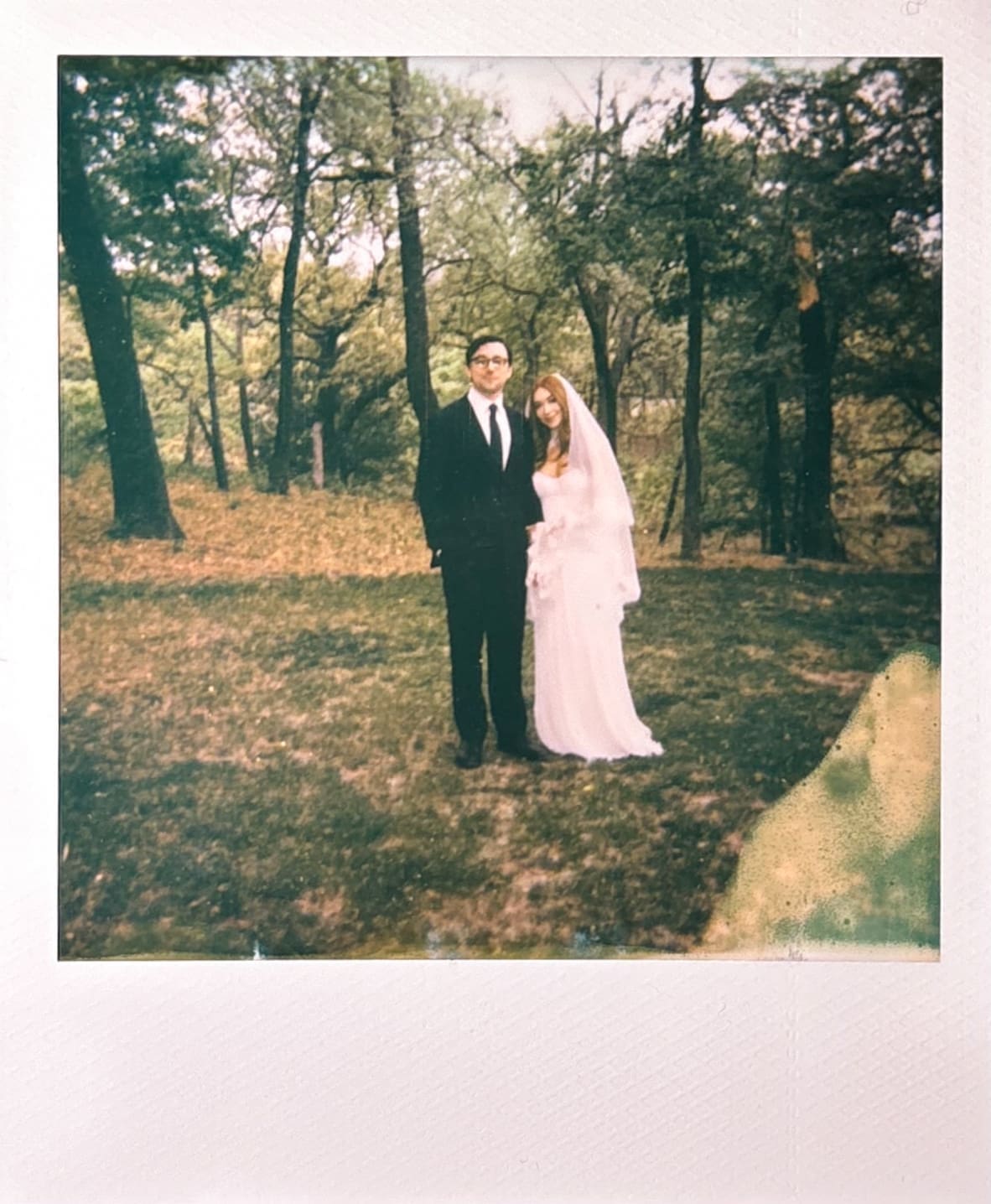 A man in a suit and a woman in a white dress stand outdoors, smiling and holding up their hands to show rings on their fingers. Trees and greenery are visible in the background.