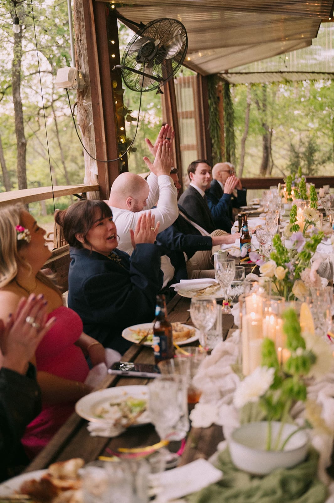 A group of people sit at a long table set for a meal, clapping and celebrating at an outdoor event decorated with flowers and candles at An Intimate Backyard Wedding in Dallas