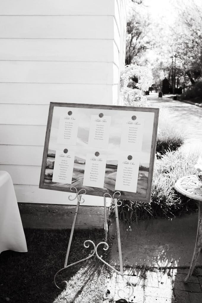 A framed seating chart displayed on a metal stand outdoors near a white building, with sunlight and garden visible in the background.