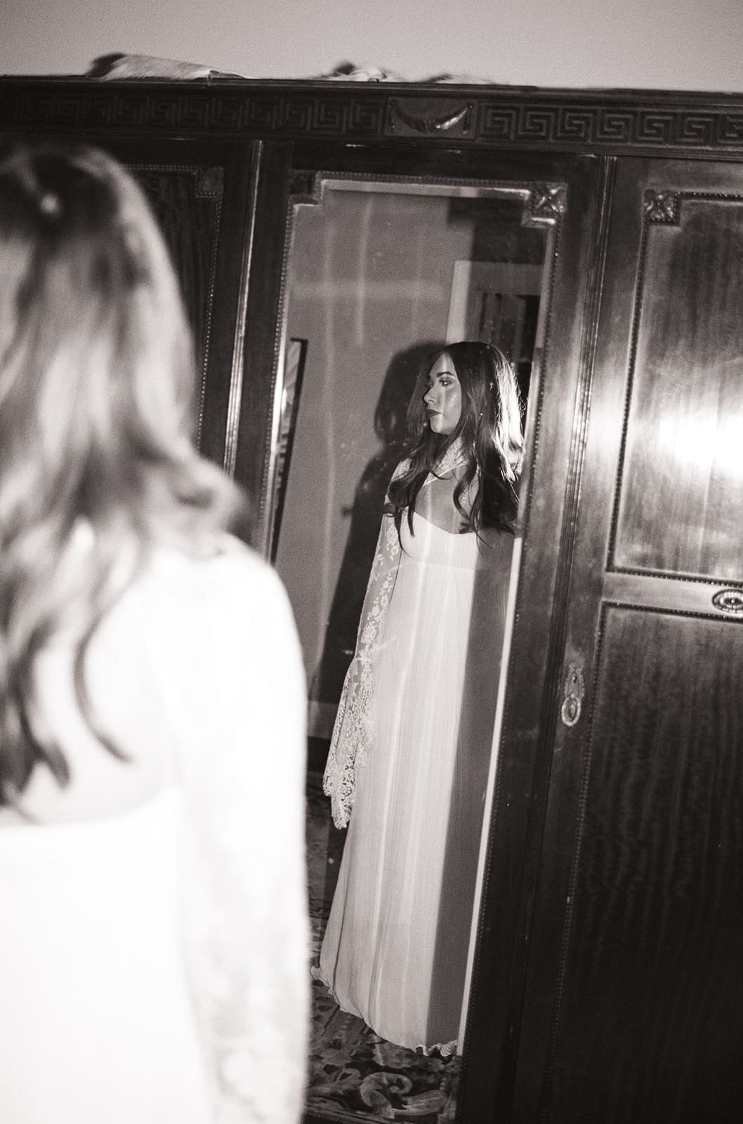 A bride in a white dress and veil stands in a room, with a woman in a floral dress smiling behind her.