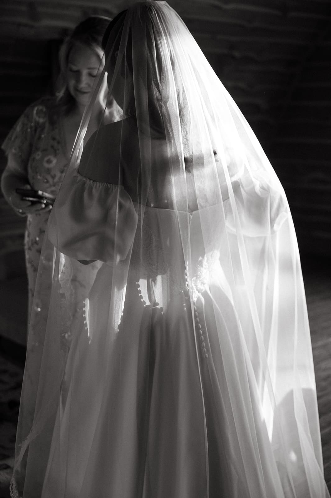 A bride in an off-shoulder wedding dress and veil stands with another woman in a dimly lit room.