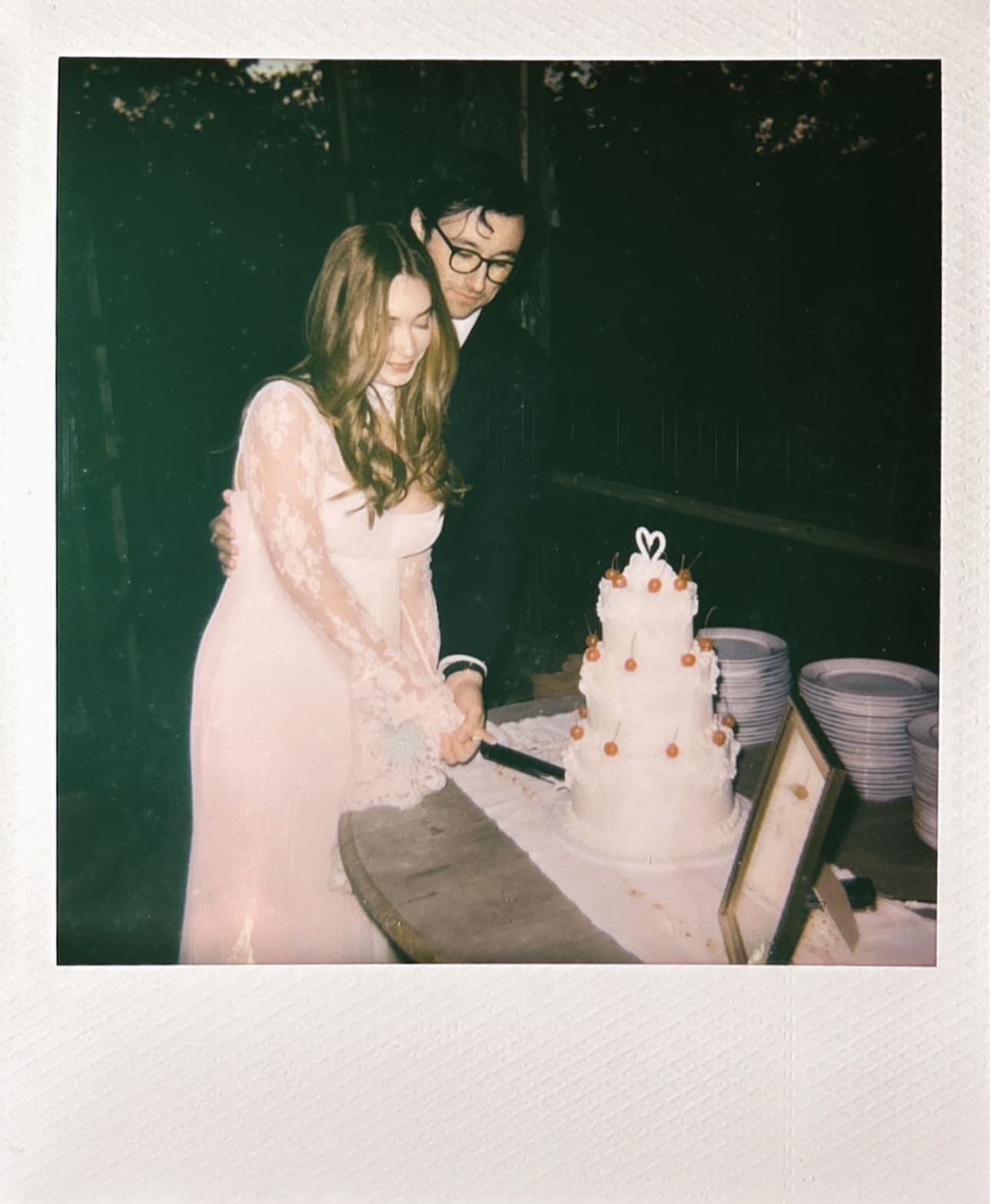 A couple in formal attire stands together cutting a tiered cake, with bowls and a framed photo on the table beside them.