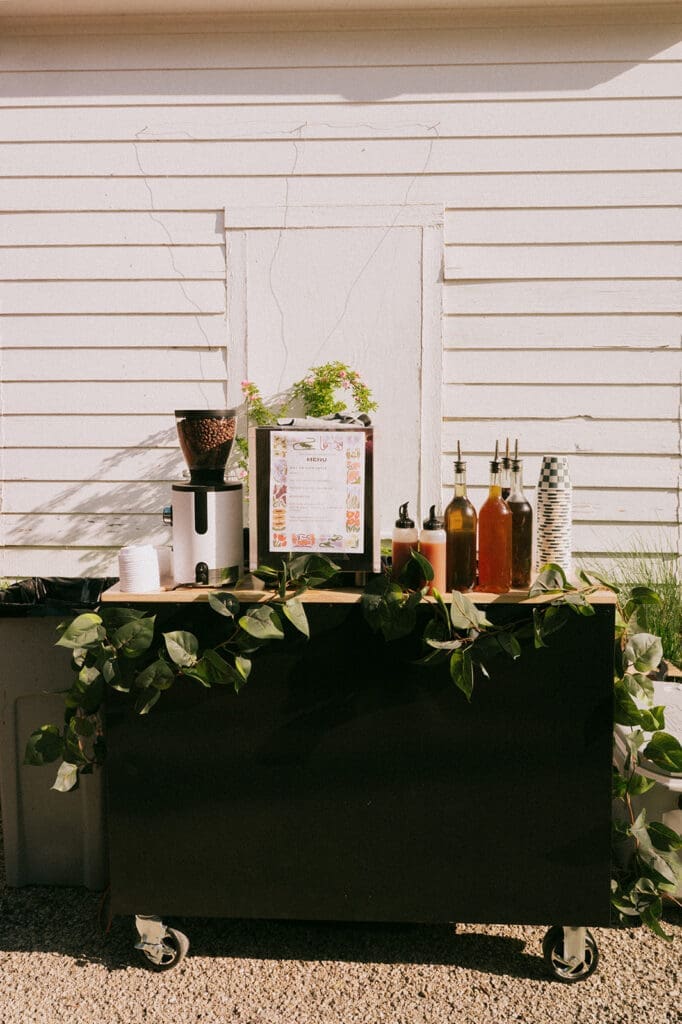 A small mobile beverage bar with various bottles, a coffee grinder, and plants is set up outdoors in front of a white wooden wall for a Weston Gardens Wedding