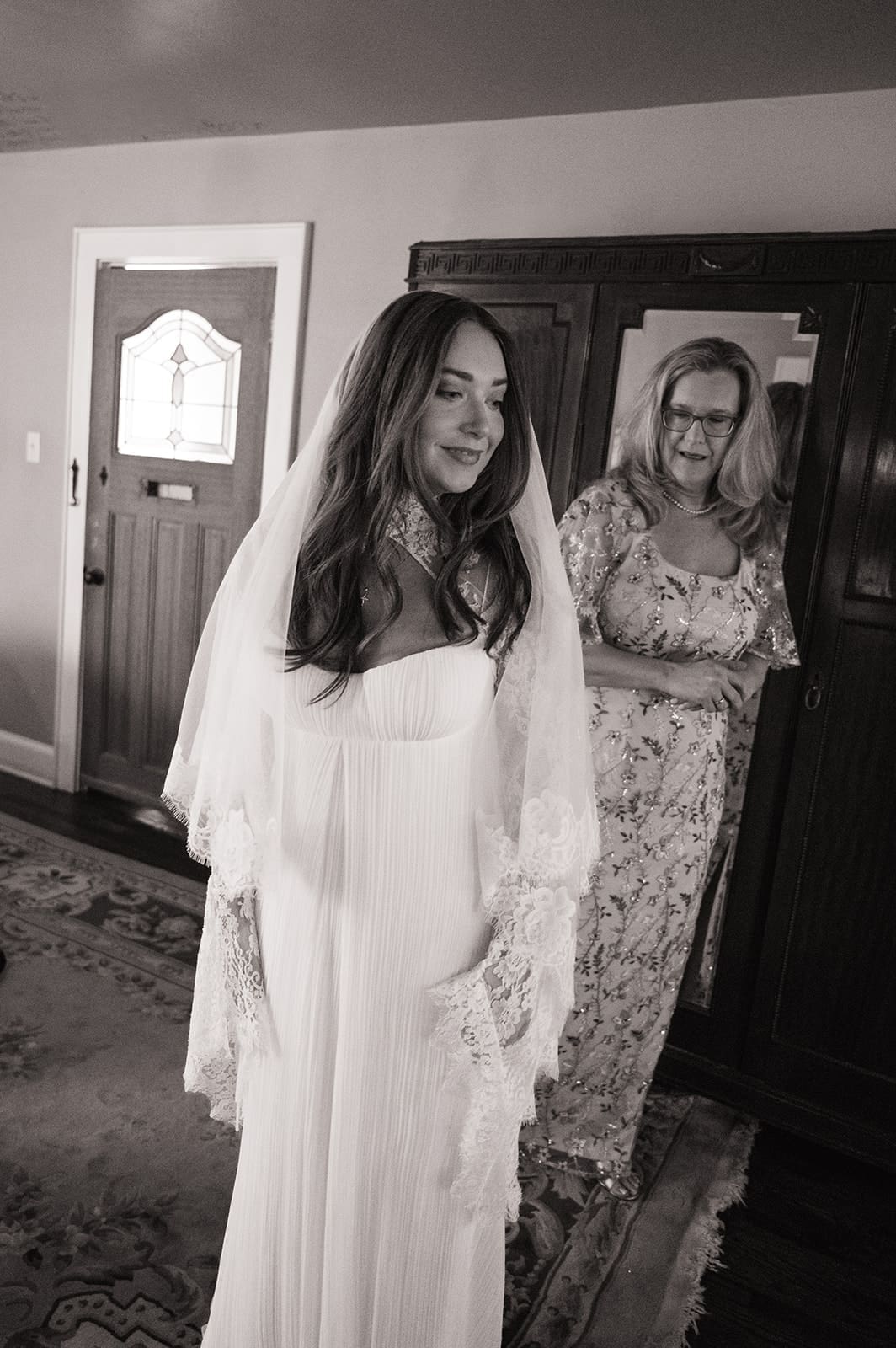 A bride in a white dress and veil stands in a room, with a woman in a floral dress smiling behind her.