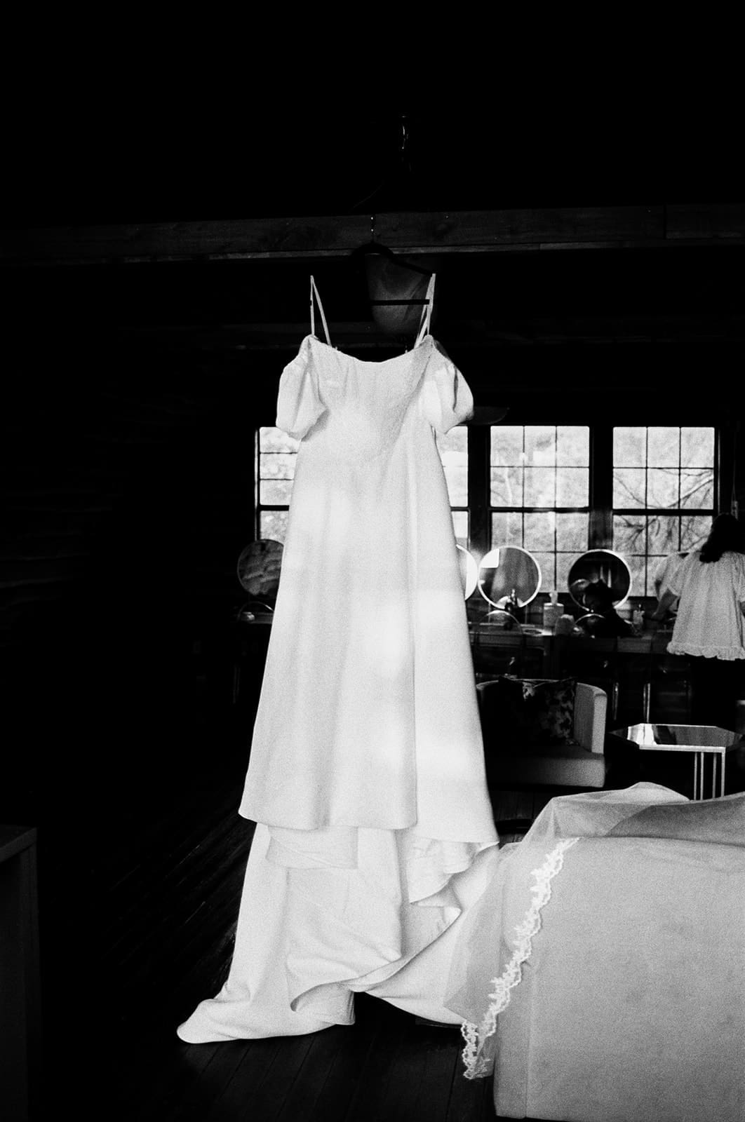 A white wedding dress on a hanger is suspended in a dimly lit room with windows and furniture visible in the background.