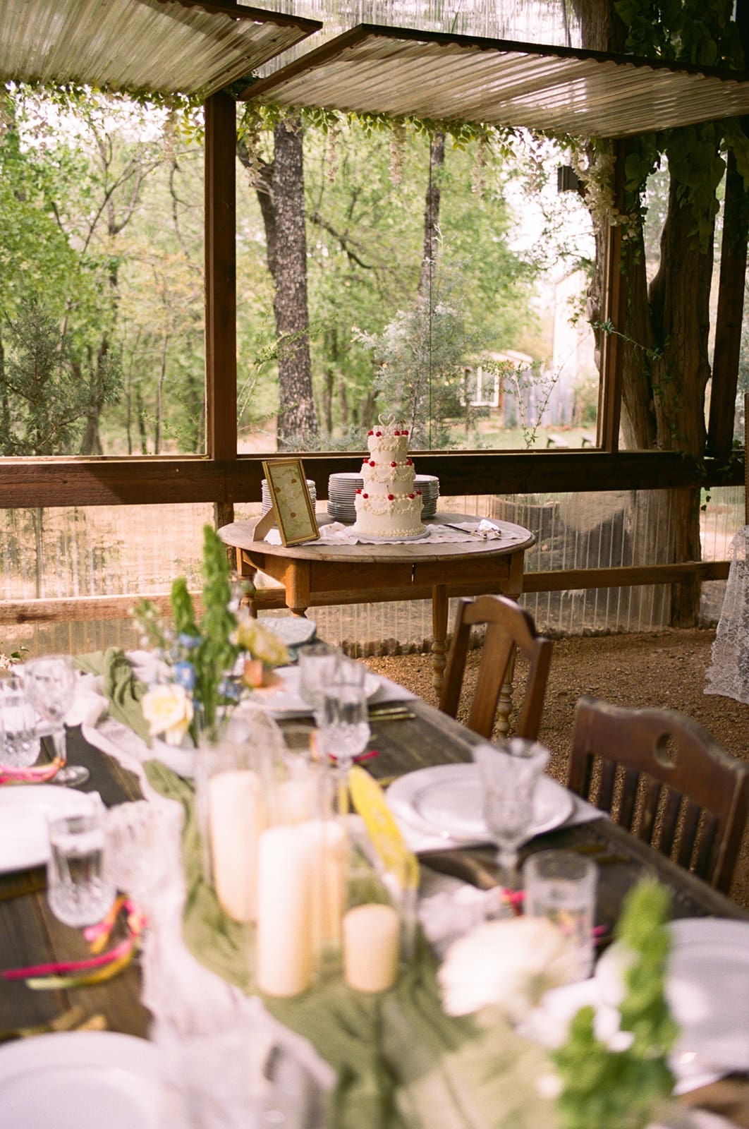 A decorated outdoor table set for a meal, with candles, glassware, and greenery, and a tiered cake displayed on a separate table in the background at An Intimate Backyard Wedding in Dallas