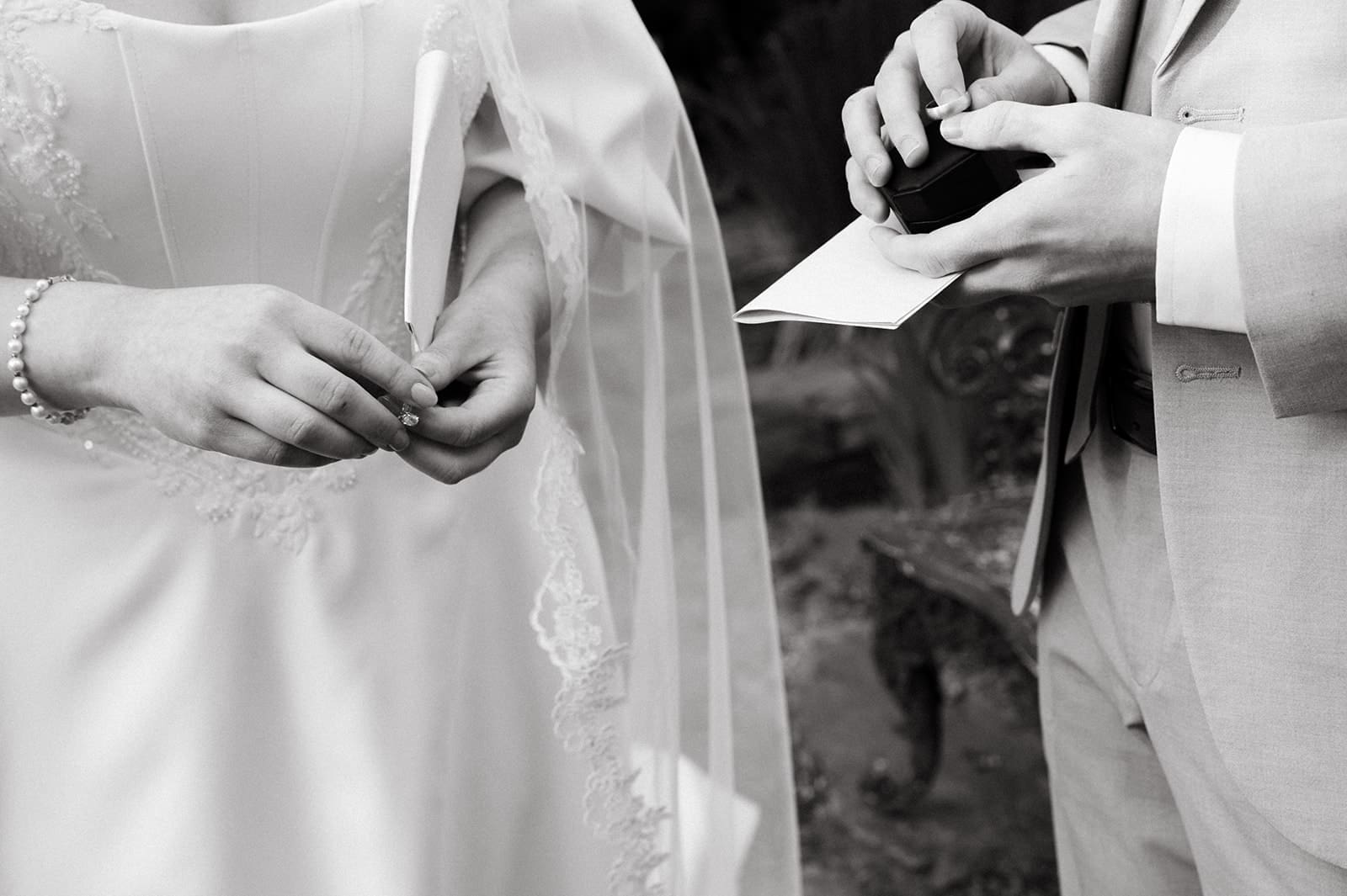 Black and white photo of a bride and groom exchanging rings and holding envelopes during a wedding ceremony, shown from the shoulders down.