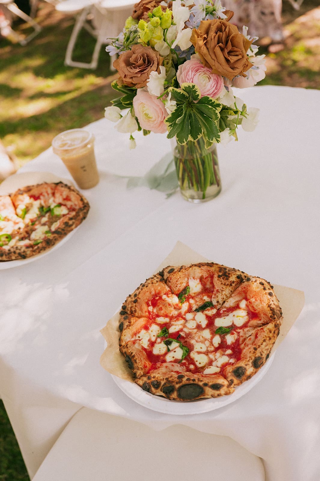 A table with two pizzas, an iced coffee, and a vase of mixed flowers set outdoors on a white tablecloth for a Weston Gardens Wedding 