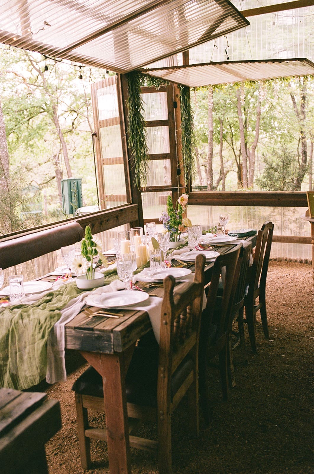 A decorated outdoor table set for a meal, with candles, glassware, and greenery, and a tiered cake displayed on a separate table in the background at An Intimate Backyard Wedding in Dallas