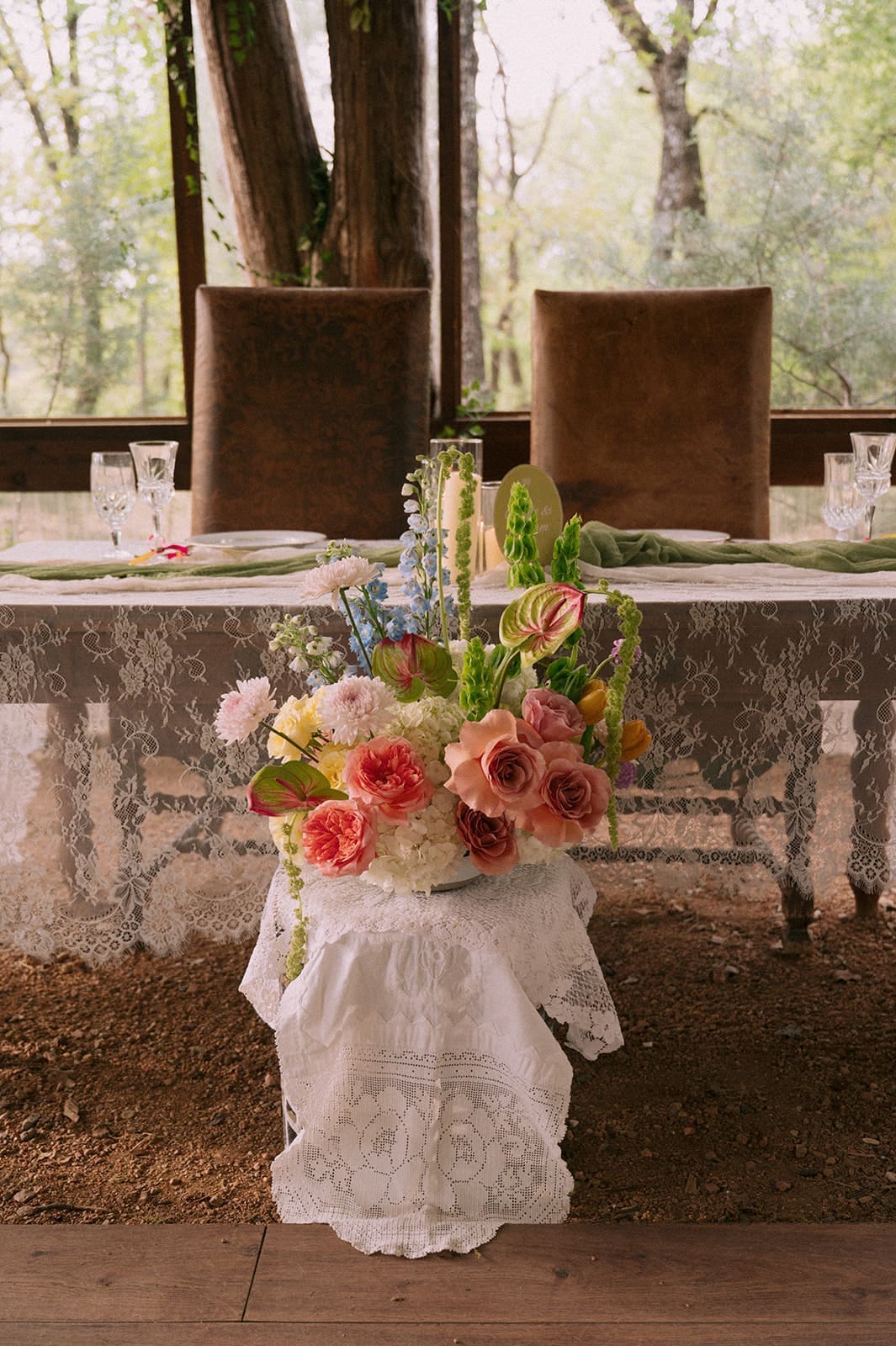 A decorated outdoor table set for a meal, with candles, glassware, and greenery, and a tiered cake displayed on a separate table in the background at An Intimate Backyard Wedding in Dallas