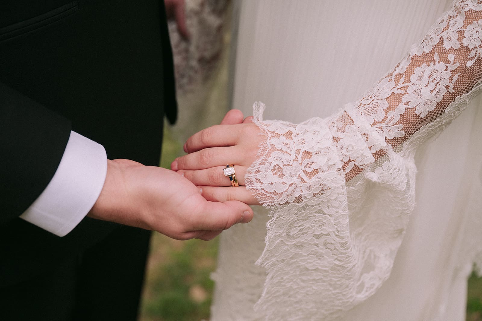 Close-up of two people holding hands, one wearing a black suit and the other in a white lace dress with a visible ring on their finger.