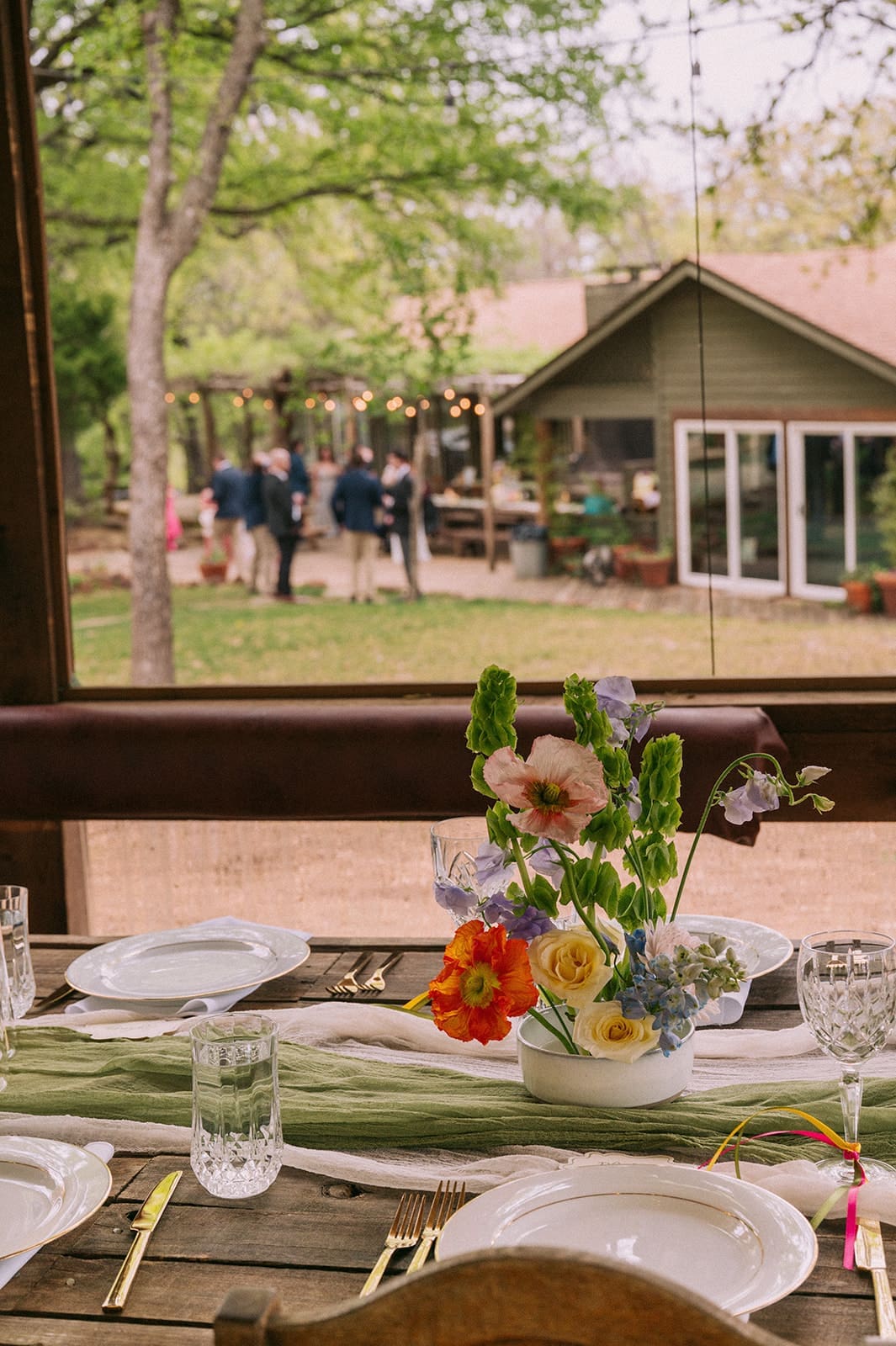A decorated outdoor table set for a meal, with candles, glassware, and greenery, and a tiered cake displayed on a separate table in the background at An Intimate Backyard Wedding in Dallas