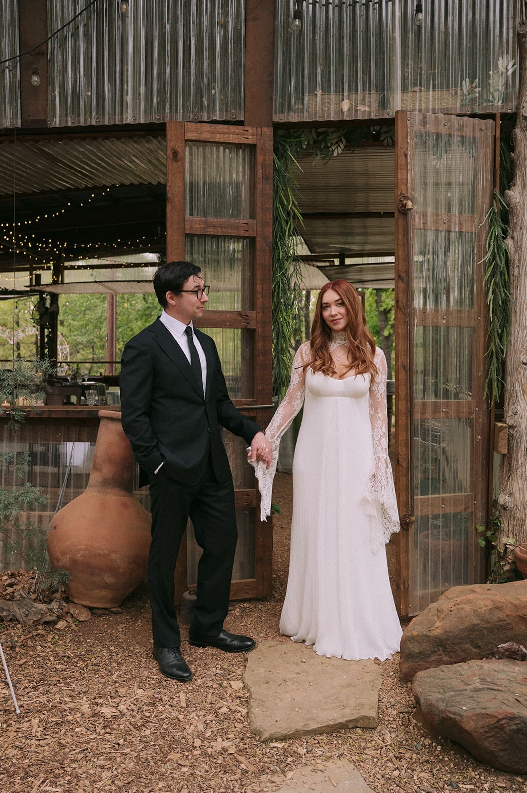 A groom in a black suit kisses the hand of a bride in a white dress as they stand outside a rustic, wooden and glass building.