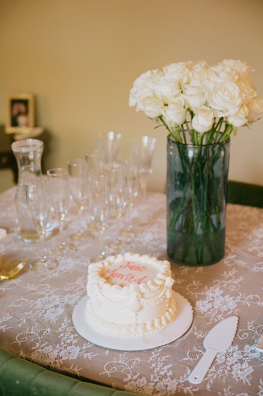 A small frosted cake, a bouquet of white roses in a vase, several empty glasses, and a cake server are arranged on a lace-covered table.