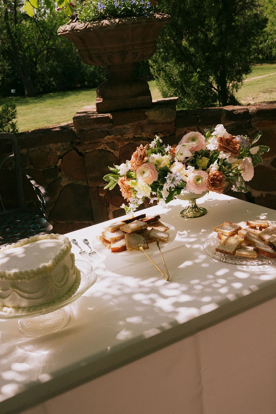 A table set outdoors with a white cake, a plate of cookies, sandwiches, and a floral arrangement, partially shaded by tree branches.