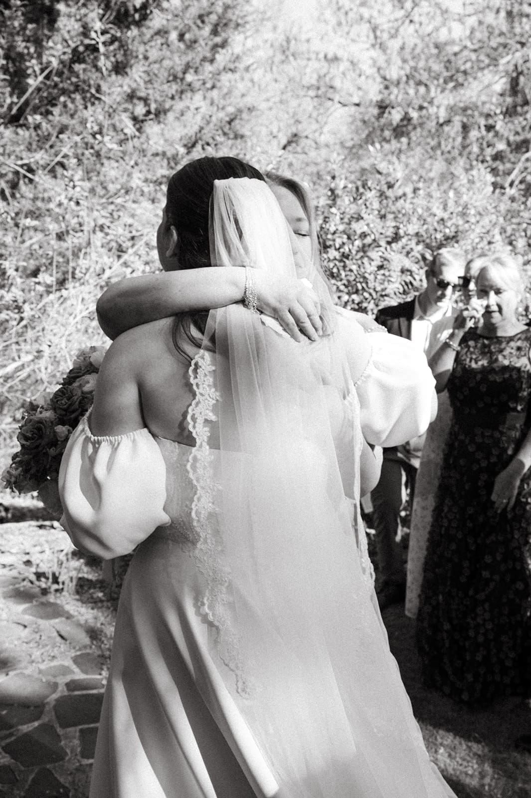 A bride in a veil and off-the-shoulder dress hugs another woman outdoors while guests watch in the background.