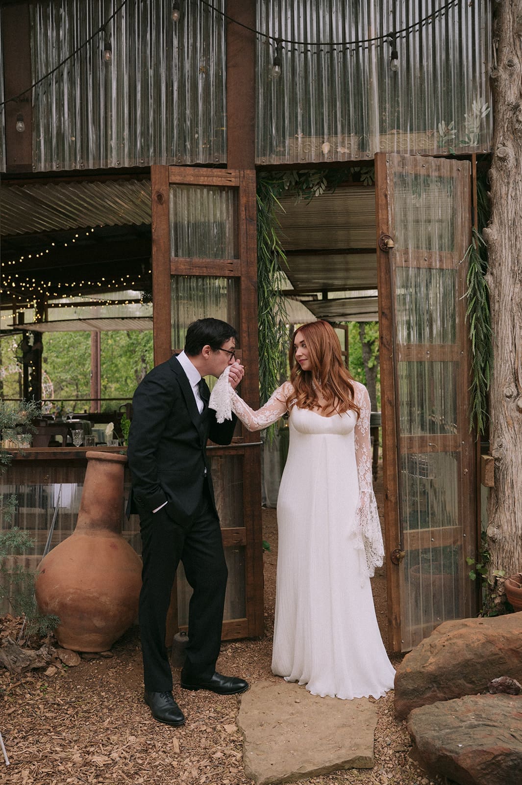 A groom in a black suit kisses the hand of a bride in a white dress as they stand outside a rustic, wooden and glass building.