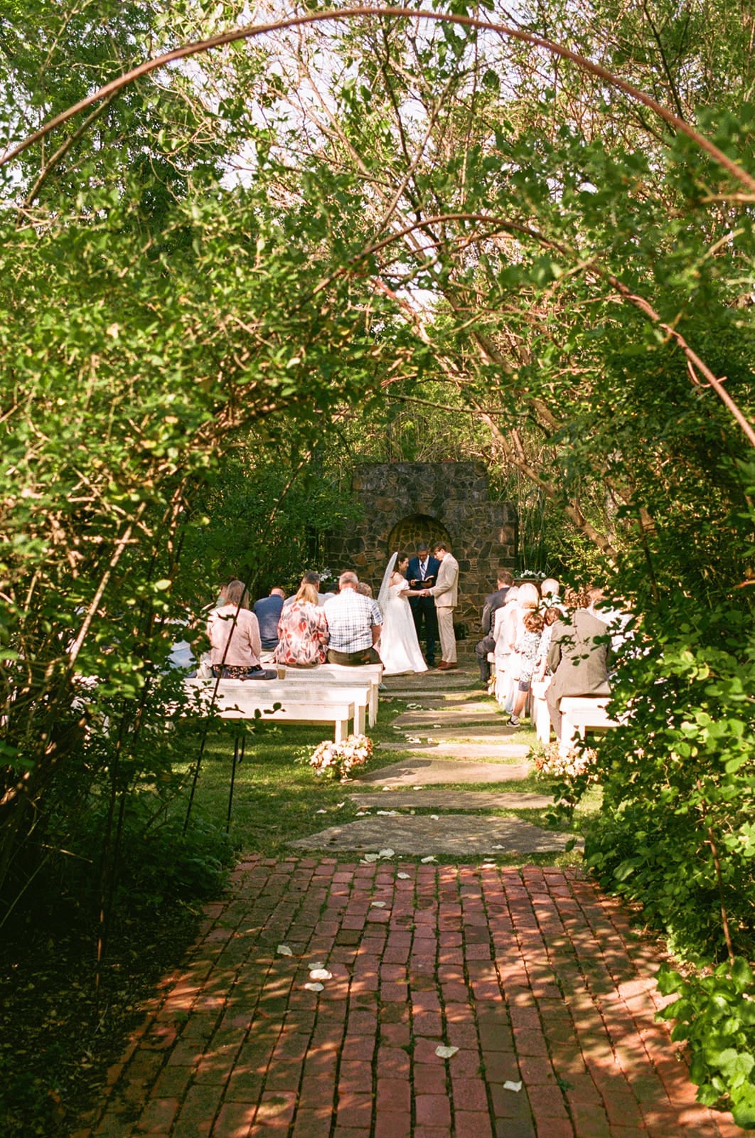 A bride and groom stand at the altar during an outdoor wedding ceremony surrounded by guests seated on benches under leafy trees.
