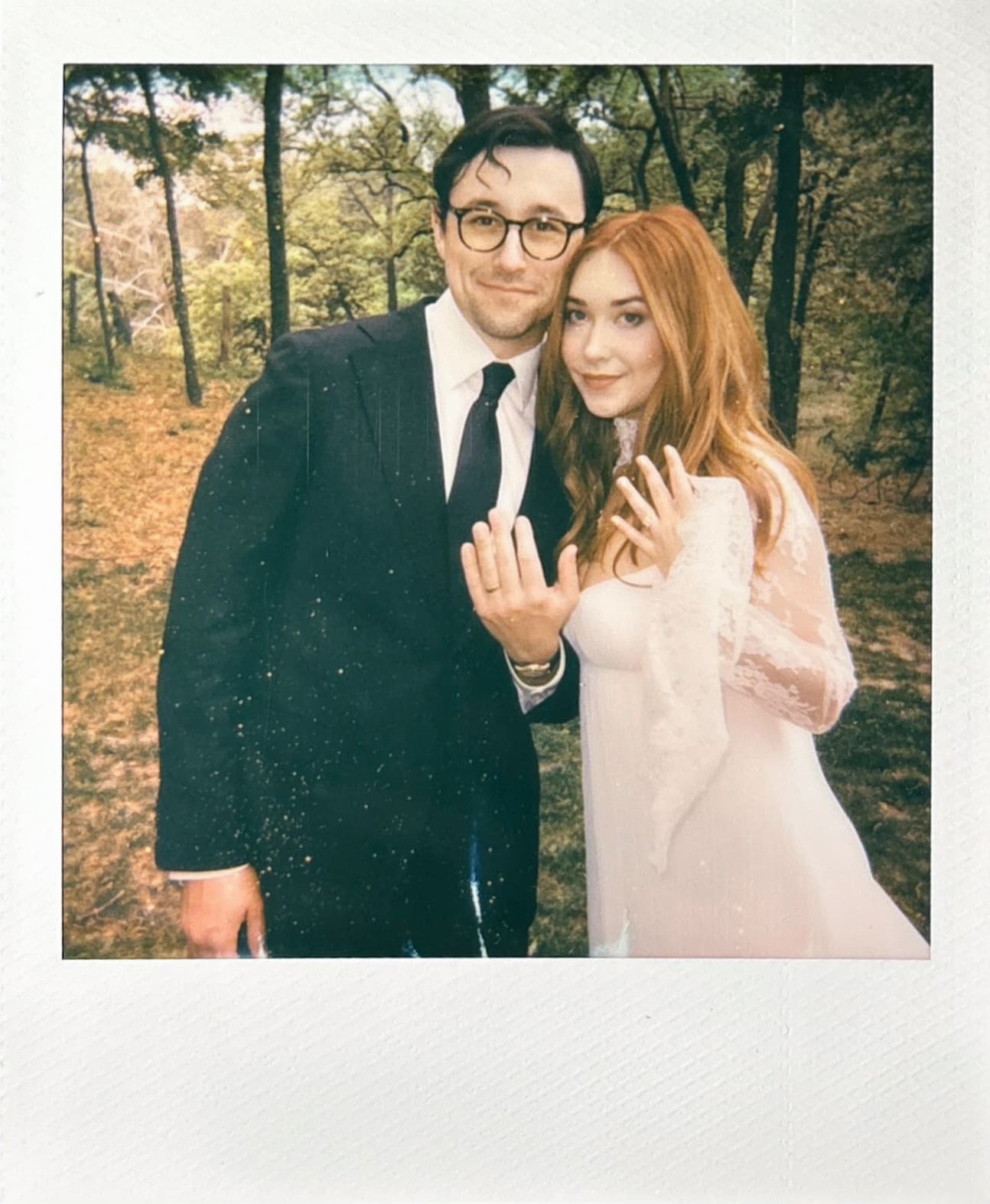 A man in a suit and a woman in a white dress stand outdoors, smiling and holding up their hands to show rings on their fingers. Trees and greenery are visible in the background.
