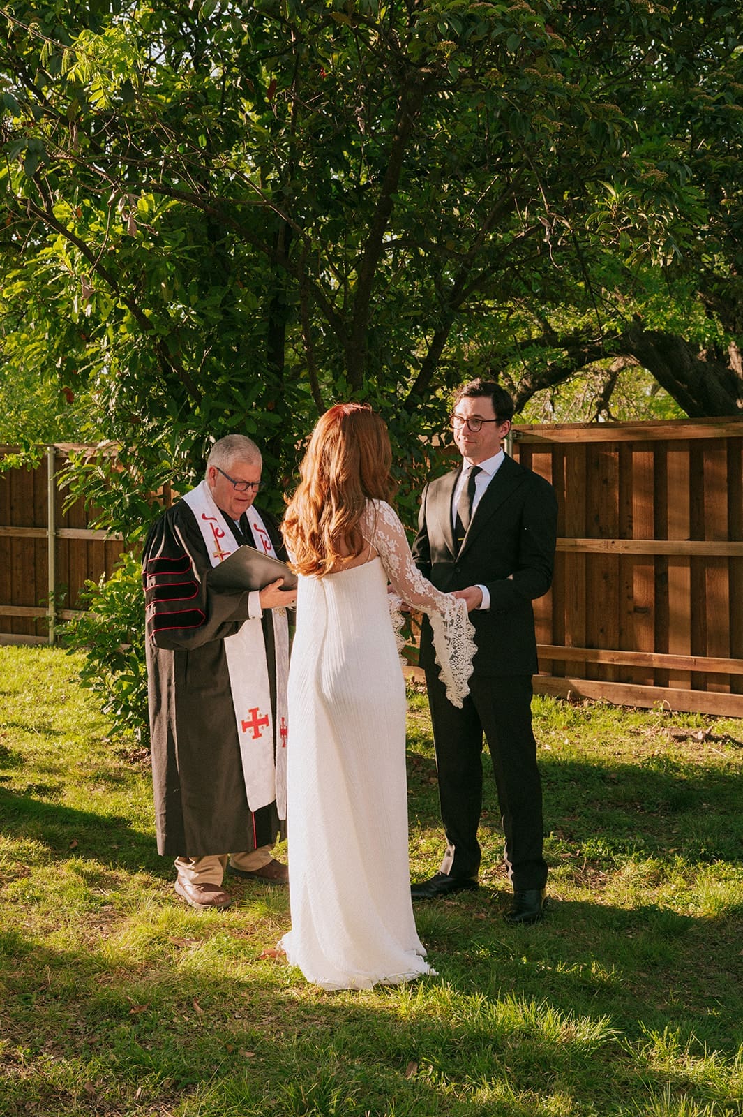 A bride and groom stand facing each other outdoors, holding hands, with an officiant reading between them during a wedding ceremony at An Intimate Backyard Wedding in Dallas