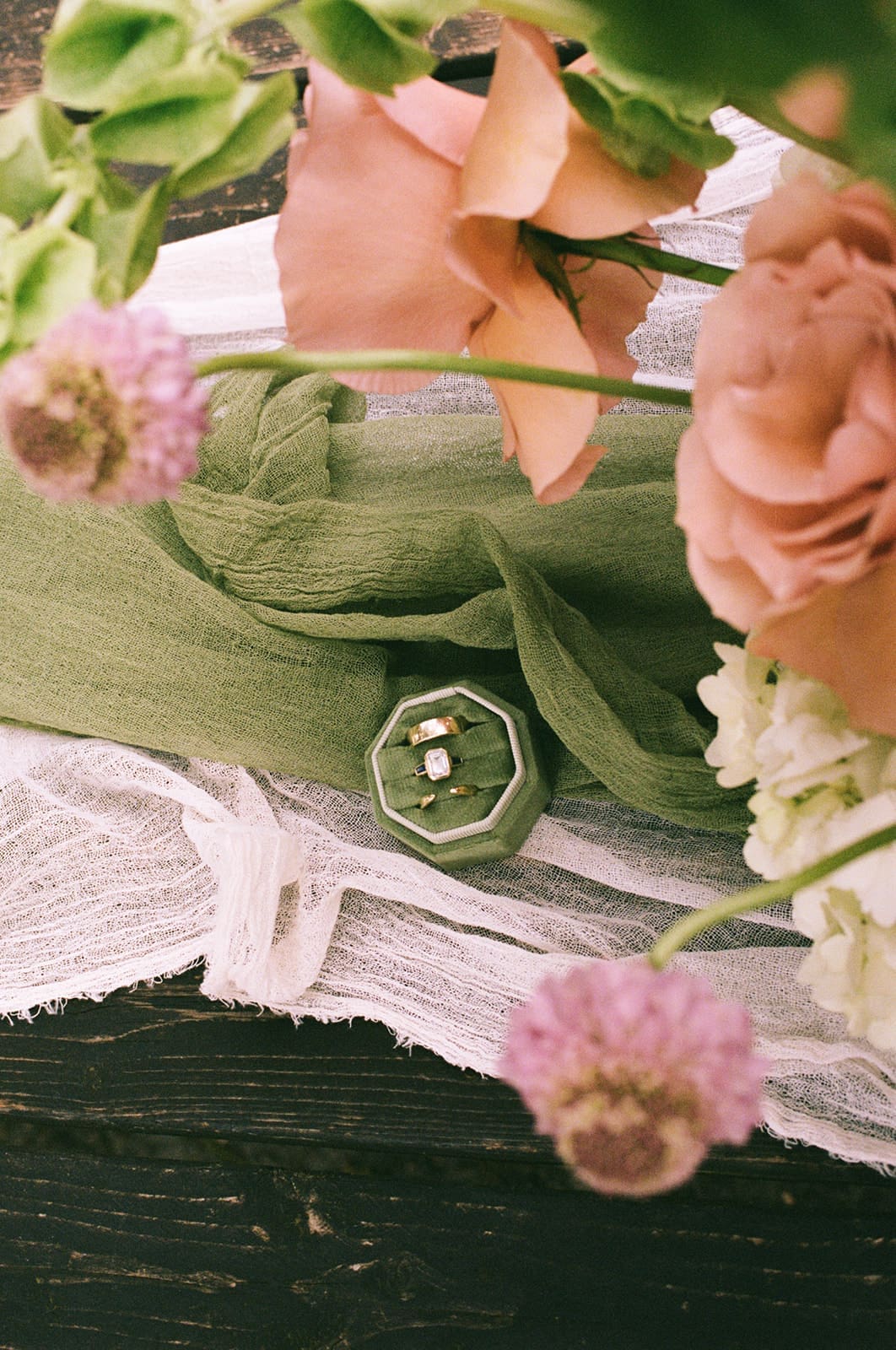A green ring box with a diamond ring inside is placed on a white cloth, surrounded by pink and peach flowers.