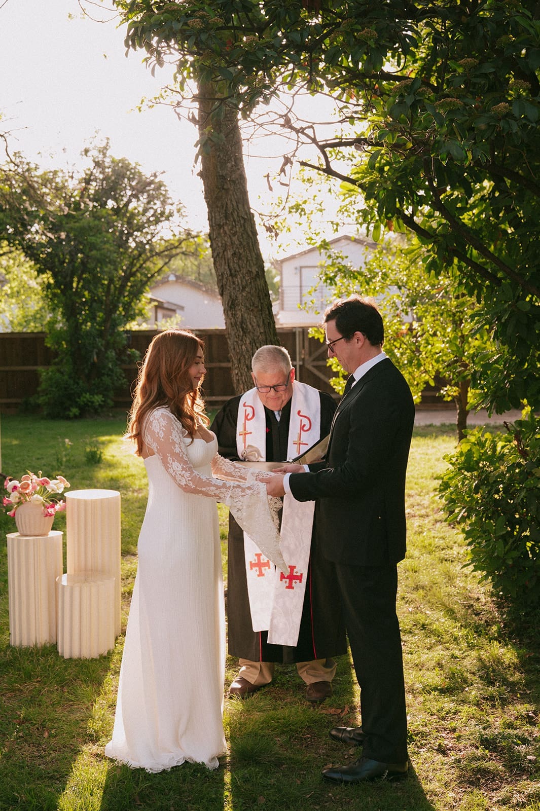 A bride and groom stand facing each other outdoors, holding hands, with an officiant reading between them during a wedding ceremony at An Intimate Backyard Wedding in Dallas