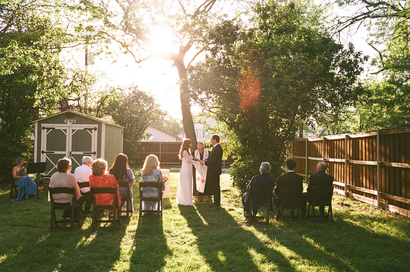 A bride and groom stand facing each other outdoors, holding hands, with an officiant reading between them during a wedding ceremony at An Intimate Backyard Wedding in Dallas