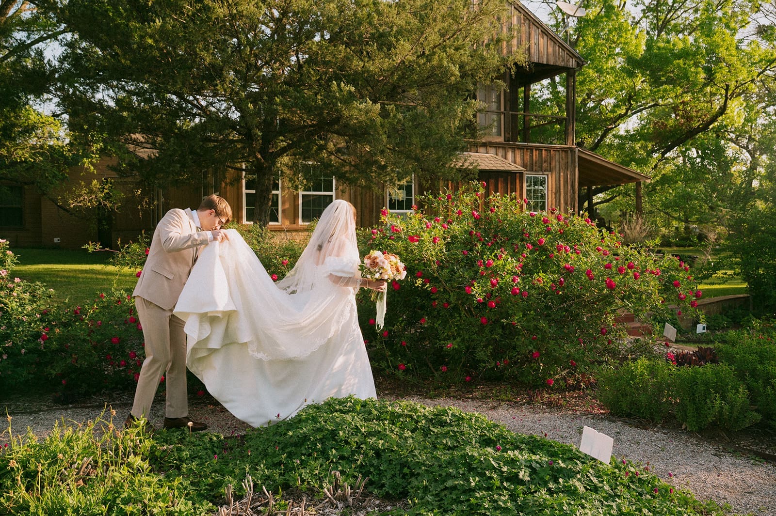 A bride and groom stand outdoors in front of a rustic wooden house, with the groom holding the bride's dress train as they walk through a garden for a weston gardens wedding