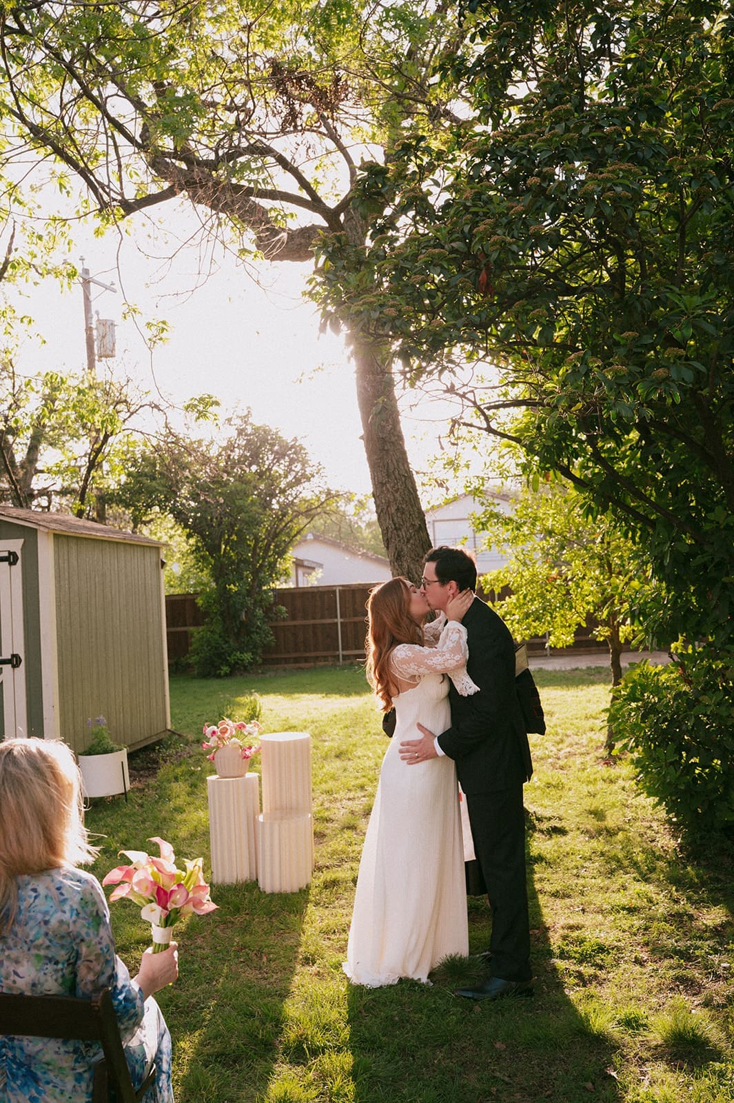 A bride and groom stand facing each other outdoors, holding hands, with an officiant reading between them during a wedding ceremony at An Intimate Backyard Wedding in Dallas