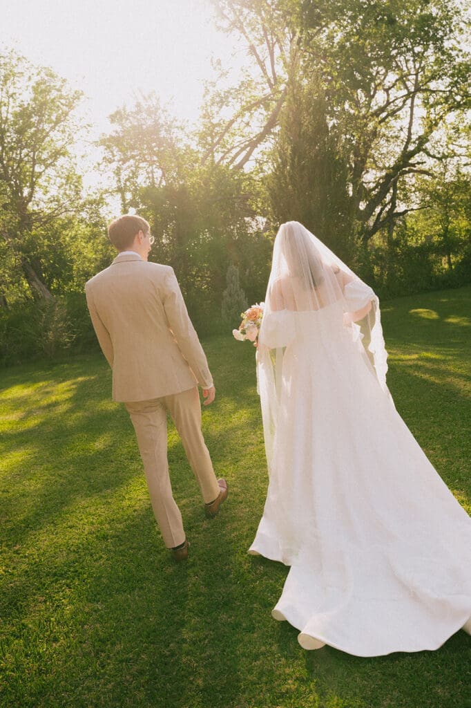 A bride in a white dress and veil walks beside a groom in a beige suit on a grassy lawn with sunlight filtering through trees for a Weston Gardens wedding