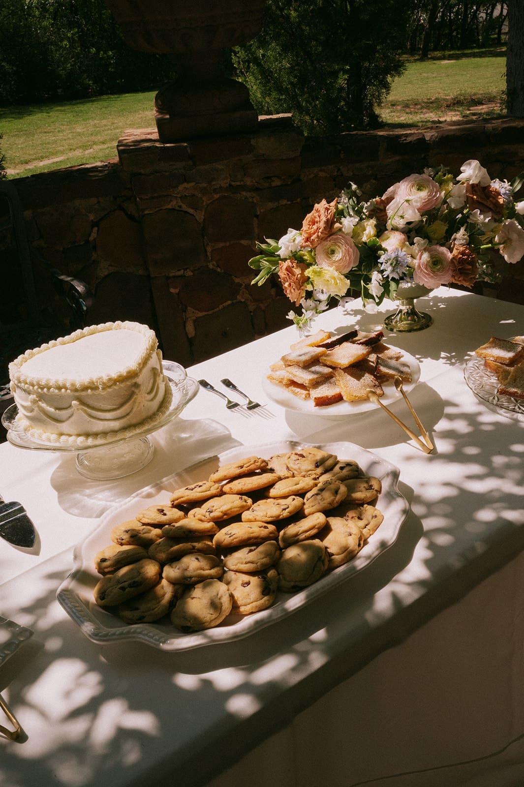 A table set outdoors with a white cake, a plate of cookies, sandwiches, and a floral arrangement, partially shaded by tree branches.