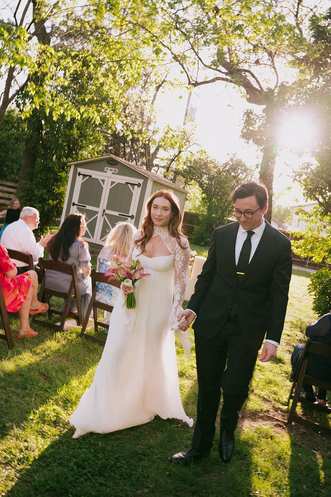 A bride and groom stand facing each other outdoors, holding hands, with an officiant reading between them during a wedding ceremony at An Intimate Backyard Wedding in Dallas