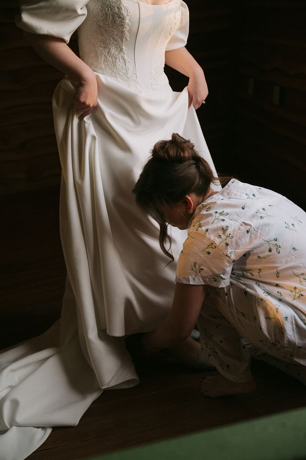 A woman in a white wedding dress stands while another person kneels to adjust the dress hem.