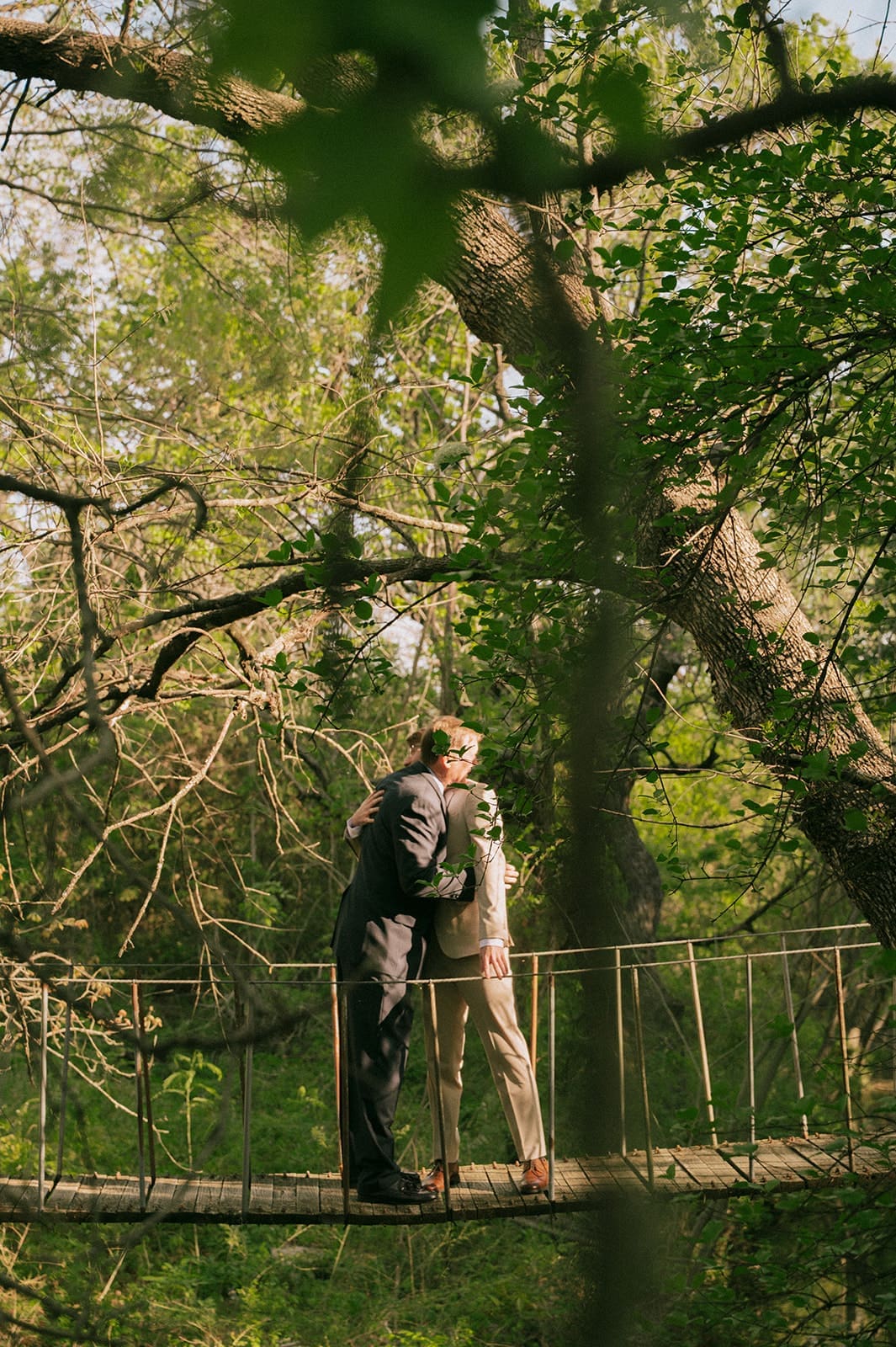 Two people stand close together on a narrow rope bridge in a forested area, surrounded by green trees and foliage.