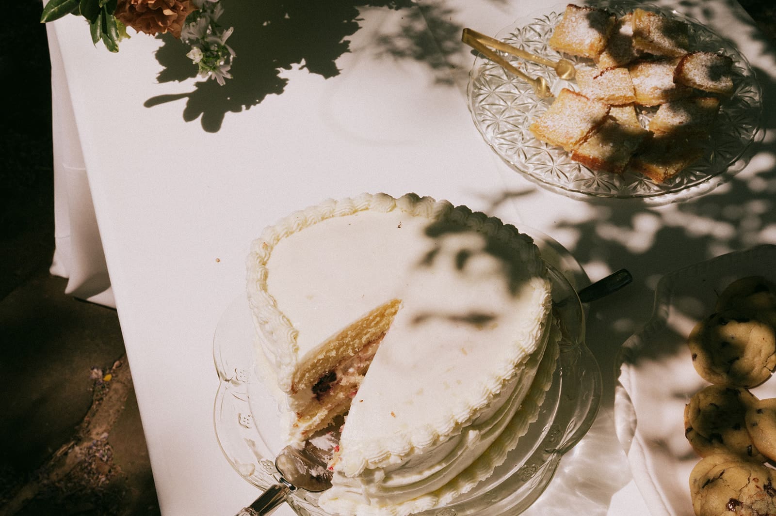 A round cake with white frosting and a missing slice sits on a table next to a plate of pastries and some cookies, partially shaded by tree branches.