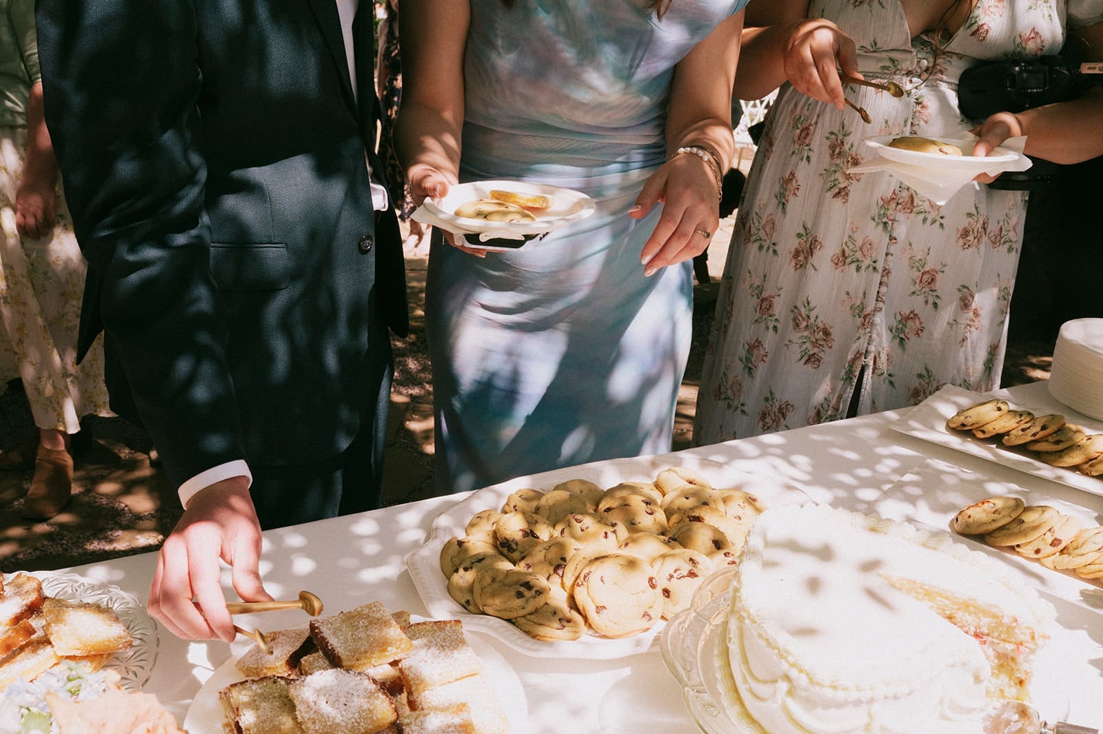 Three people in formal attire serve themselves dessert from a table with cookies, cake, and other pastries at an outdoor event.