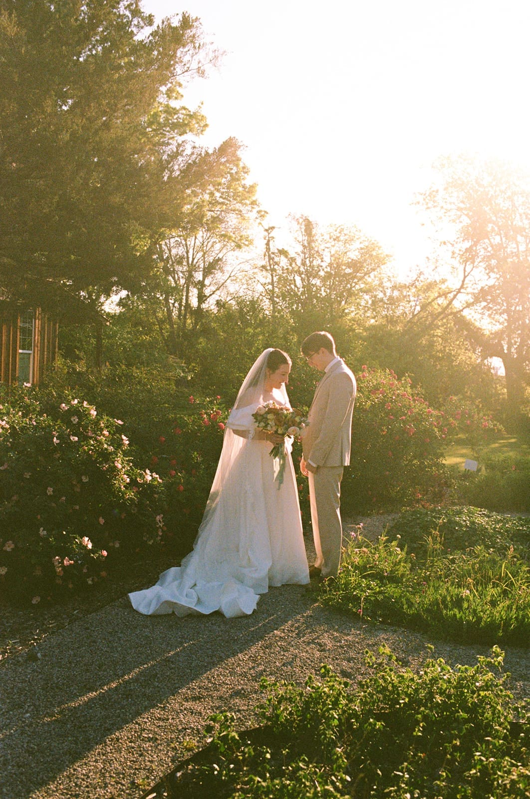 A bride in a white dress and veil stands beside a groom in a beige suit on a grassy lawn with sunlight filtering through trees for a Weston Gardens wedding