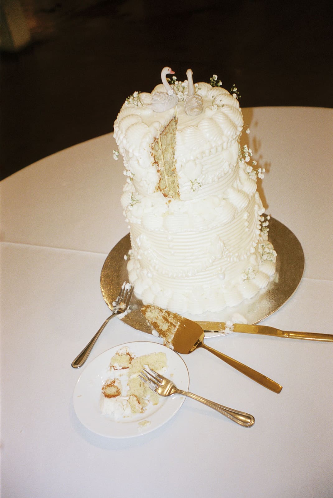 A two-tier white cake with decorative frosting sits on a table, partially sliced, with serving utensils and a small plate containing a piece of cake nearby.