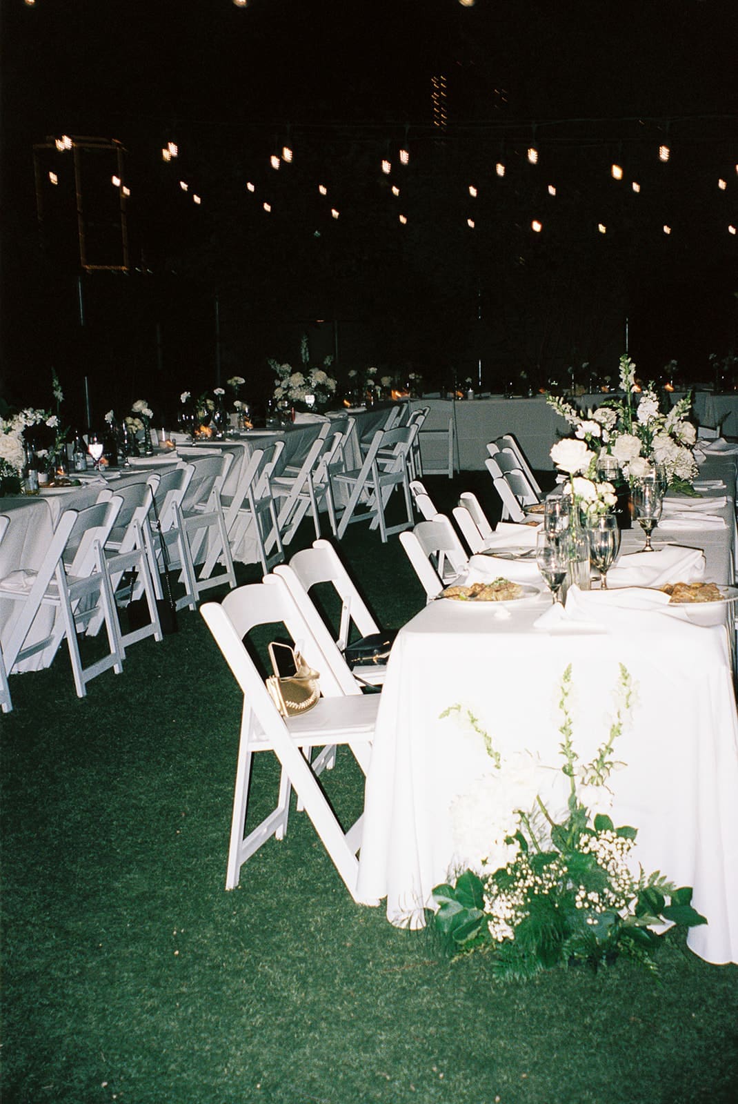White tables and chairs arranged outdoors on grass, decorated with flower centerpieces and string lights overhead, prepared for an event at a small wedding venue in dallas tx
