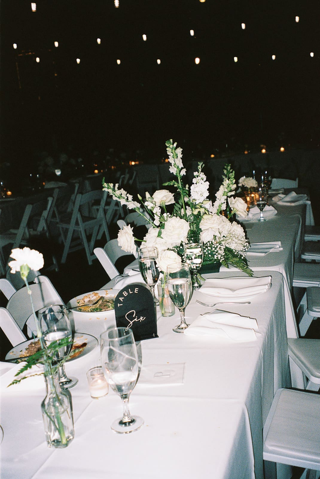 A group of people in formal attire gather and mingle outdoors under string lights, surrounded by trees and greenery at a small wedding venue in dallas tx