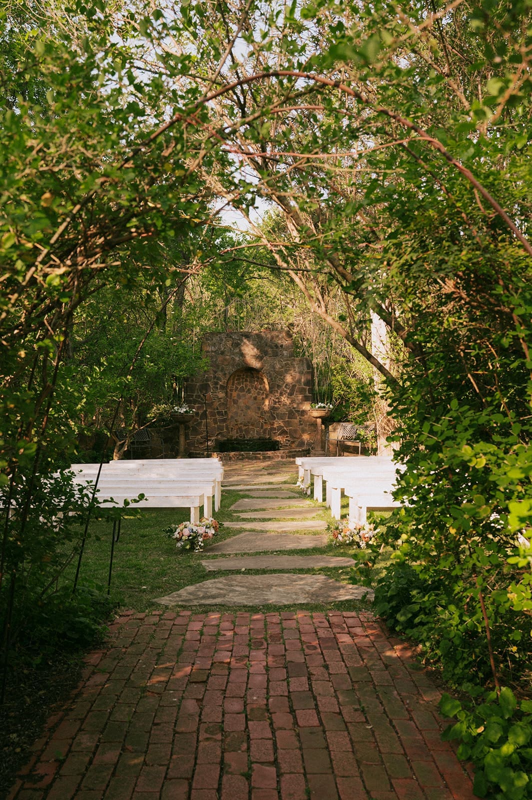 A brick pathway leads through an arch of leafy trees to an outdoor stone structure, with white benches arranged on either side at a Small Wedding Venue in Dallas, TX